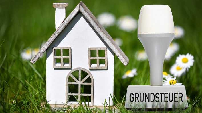 Fotomontage: Miniaturhaus im Grünen und Stempel mit Aufschrift Grundsteuer, Foto: IMAGO / Christian Ohde